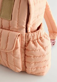 Pink padded backpack featuring a quilted texture, elastic side pocket, and flap pocket with a fabric strap detail.