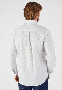 Chemise à boutons pour homme en blanc avec un motif noir répétitif. Dotée de manches longues et d'un col classique, avec une pièce de dos et un détail de pli.