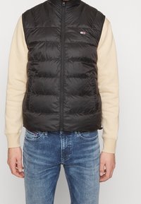 Czarna pikowana kamizelka z wysokim kołnierzem, zapinana na zamek, z poziomymi przeszyciami, noszona na kremowej bluzce z długim rękawem. Delikatne logo na piersi.