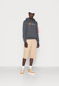 Felpa grigia con cappuccio e logo arancione, pantaloni cargo beige e sneakers alte color beige. Il modello indossa un cappello bianco e si trova davanti a uno sfondo semplice.
