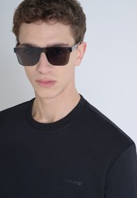 T-shirt nera con scollo rotondo e maniche corte, con un logo discreto sul petto. Occhiali da sole scuri e rettangolari con un accento metallico.