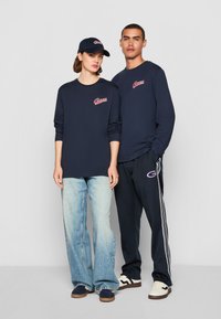 Marine langermet T-skjorte med brodert logo, kombinert med lyseblå jeans og mørke joggesko; matchende marinecaps og joggebukser med hvite striper.