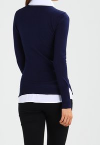 Marineblauwe gebreide trui met lange mouwen, slank ontwerp en geribde zoom, gelaagd over een witte blouse met kraag.