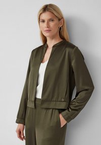Giacca in satin verde oliva con colletto rotondo, polsini elasticizzati e chiusura a due bottoni in vita. Indossata sopra un top bianco, di texture liscia.