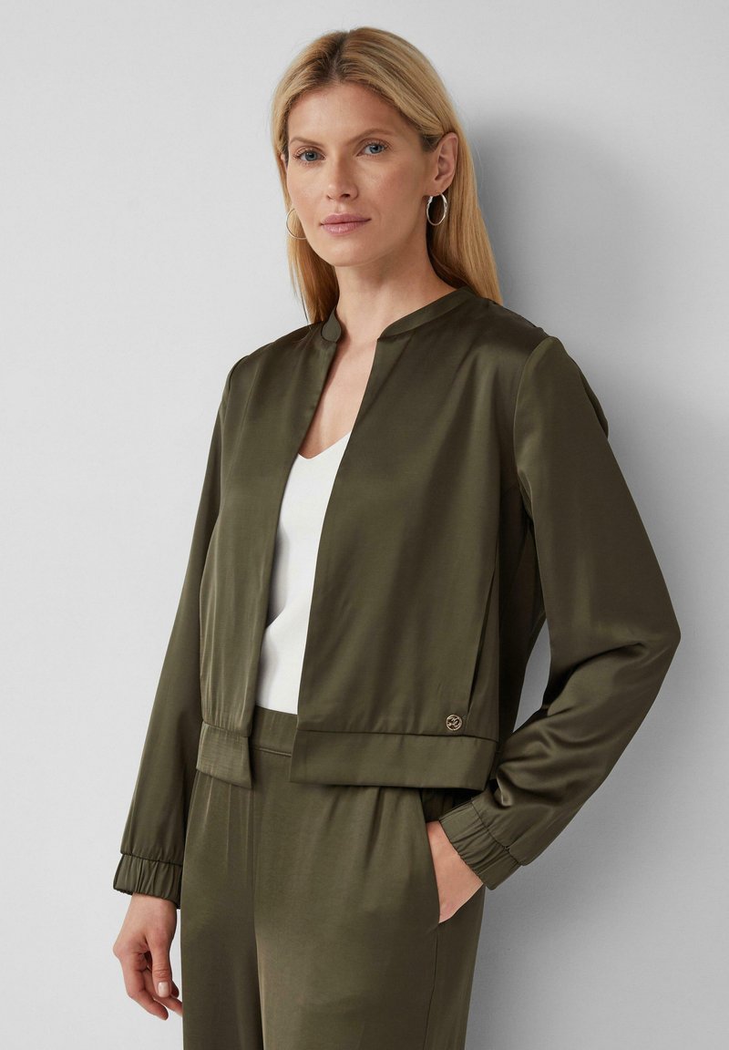 Giacca in satin verde oliva con colletto rotondo, polsini elasticizzati e chiusura a due bottoni in vita. Indossata sopra un top bianco, di texture liscia.