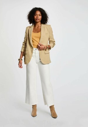 Blazer in lino beige sopra un top senape, abbinato a pantaloni bianchi a gamba larga e stivaletti beige. Accessori dorati completano il look.