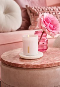 Vit ribbad ljus med en guldlapp på en rund marmorbasis, placerad på ett rosa marmorbord bredvid en rosa blomma i en glasvas.