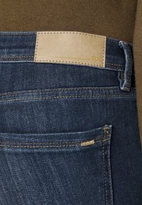 Mörkblå denimjeans har ett brunt läderpatch vid midjan, guldtråstadder och en bakficka med detalj i metallknapp.