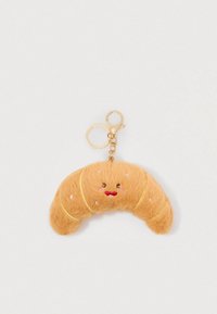Porte-clé en forme de croissant moelleux avec un visage souriant brodé et un anneau et un fermoir en or sur un fond blanc.