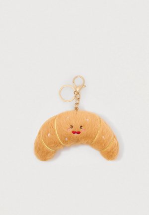Porte-clé en forme de croissant moelleux avec un visage souriant brodé et un anneau et un fermoir en or sur un fond blanc.