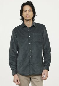 Chemise boutonnée en velours côtelé vert foncé avec une poche poitrine unique, devant boutonné et manches longues. Texture lisse et motif côtelé.