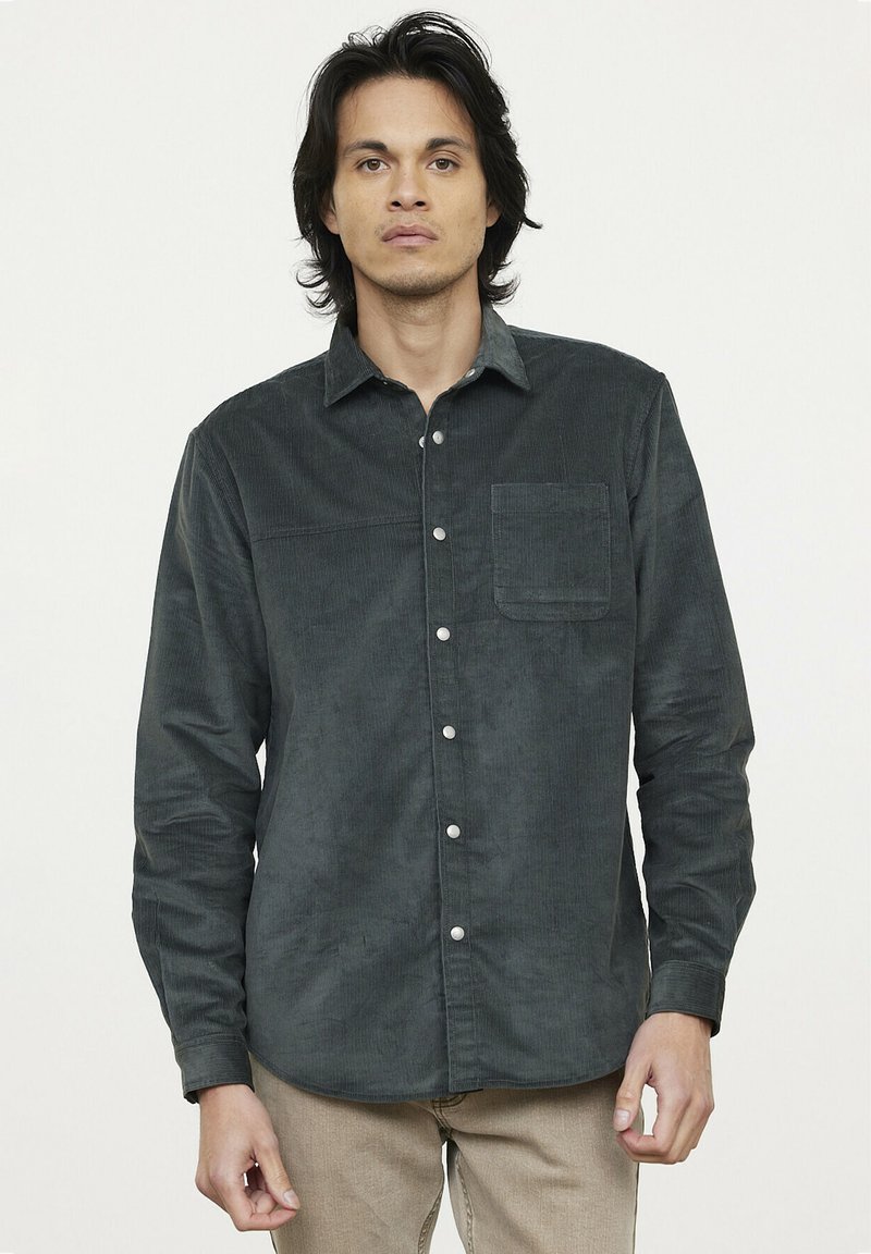 Chemise boutonnée en velours côtelé vert foncé avec une poche poitrine unique, devant boutonné et manches longues. Texture lisse et motif côtelé.