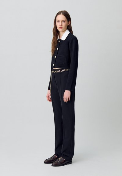 Cardigan nero corto con colletto bianco, due tasche frontali e bottoni dorati, abbinato a pantaloni neri a vita alta e mocassini marroni.