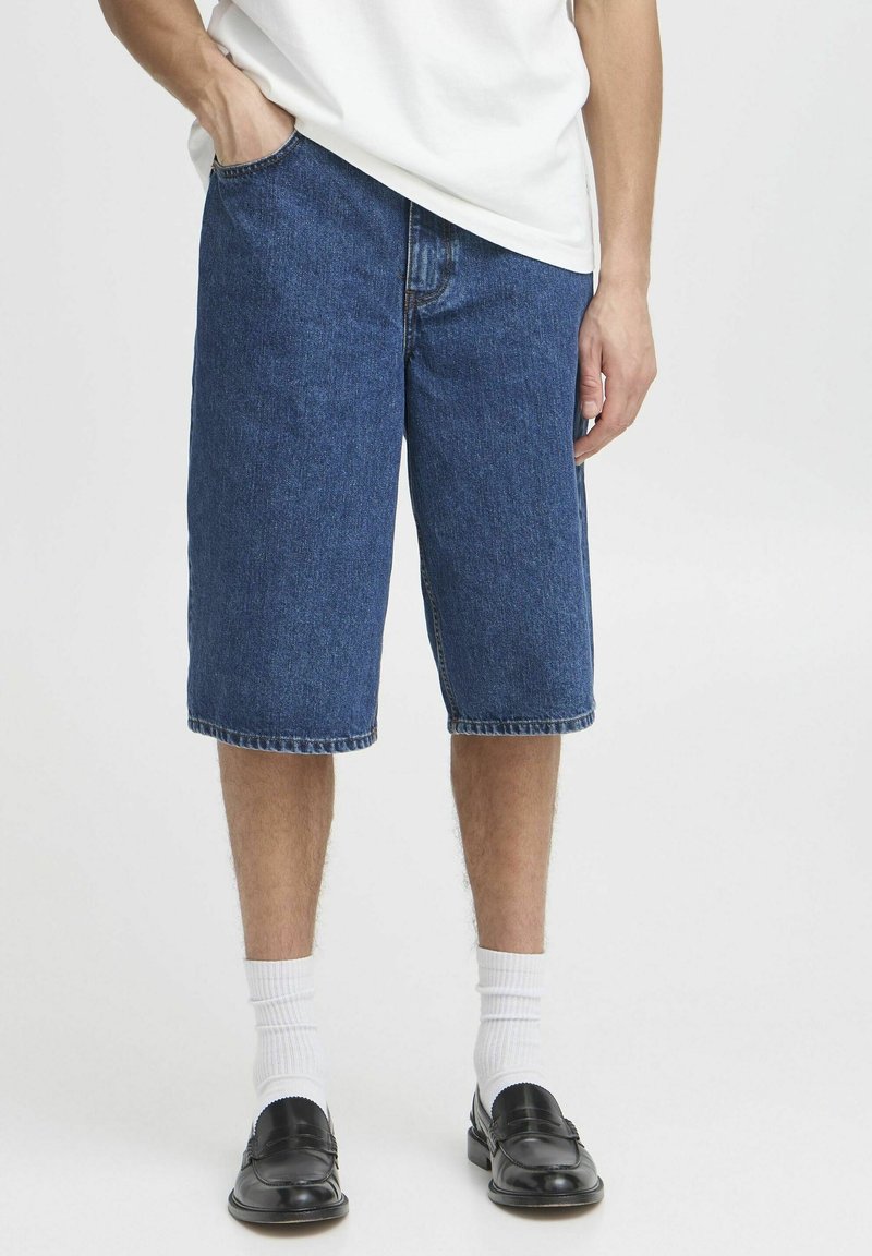 Homme portant un short en denim bleu jusqu'aux genoux, des chaussettes blanches mi-hautes, des mocassins noirs et un t-shirt blanc, avec une main dans la poche.
