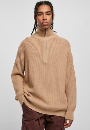 Pull en maille côtelée beige avec col montant et fermeture éclair à l'avant. Coupe oversize avec épaules tombantes et manches longues. Tissu en maille texturée.