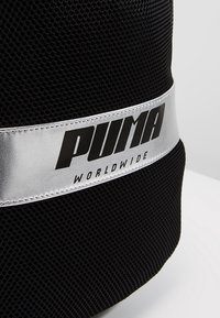 Mochila de malla negra con una banda plateada que presenta el texto "PUMA" y "WORLDWIDE" en negrita. El diseño destaca por su superficie texturizada.