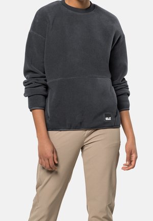 Osoba ubrana w ciemnoszarą bluzę z długim rękawem z małą naszywką z logo oraz beżowe spodnie na jednolitym tle.