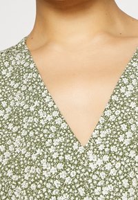 Grünes Blumenkleid mit V-Ausschnitt, verziert mit kleinen weißen Blumen auf einem strukturierten Stoff, Nahaufnahme des Halsbereichs.