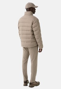 Man die met zijn rug naar de camera staat, gekleed in een beige pufferjack, bijpassende broek, een pet en chunky sneakers op een effen lichte achtergrond.