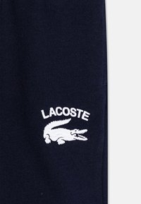 Marinblå bomullsbyxor i sweatshirtmaterial har en broderad vit Lacoste-logotyp med en krokodildesign på vänster ben. Mjuk tygtextur.