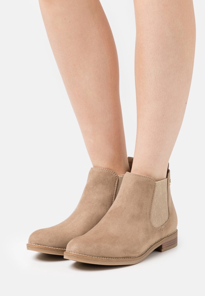 Bottines beiges en daim avec des petits talons en bois, portées sur jambes nues, avec des panneaux élastiques latéraux pour un enfilage facile.