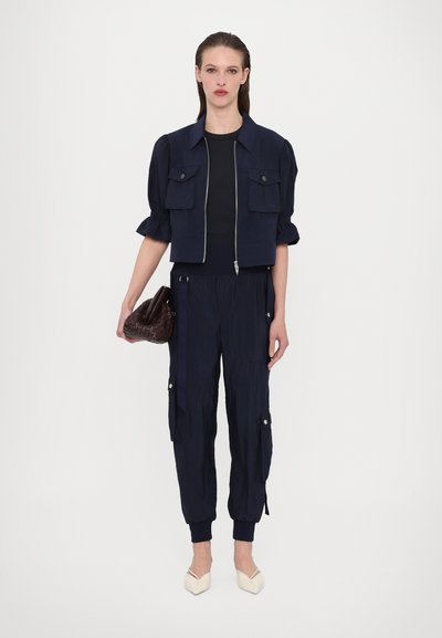 Veste courte bleu marine avec des manches bouffantes, fermeture éclair, deux poches poitrine. Pantalons cargo bleu marine assortis avec des poches latérales. Chaussures plates beige à bout pointu.