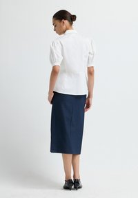 Witte blouse met pofmouwen, een getailleerde taille en een peplum-zoom, gecombineerd met een donkerblauwe kokerrok. Model draagt zwarte hakken.