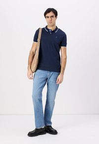 Camisa polo azul marino con acentos blancos, jeans de color azul claro y zapatos negros. El modelo lleva una bolsa de mensajero marrón claro, de pie contra un fondo blanco.