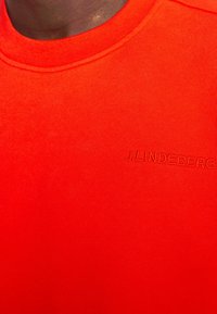 Červené bavlněné tričko s žebrovaným kulatým výstřihem a vyraženým logem "J.LINDEBERG" na levé straně, s hladkou a jednotnou texturou.