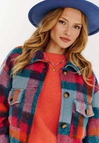 Károvaná vlnená bunda v modrej, červenej a tyrkysovej farbe s veľkými gombíkmi a náprsními vreckami. V kombinácii s oranžovým sveterom a modrým klobúkom s širokým okrajom.