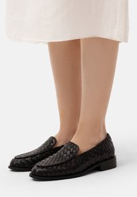Schwarze geflochtene Loafers mit flachem Absatz, die mit einem glatten Lederbesatz und einem runden Zehendesign ausgestattet sind, auf nackten Füßen vor einem hellen Hintergrund präsentiert.
