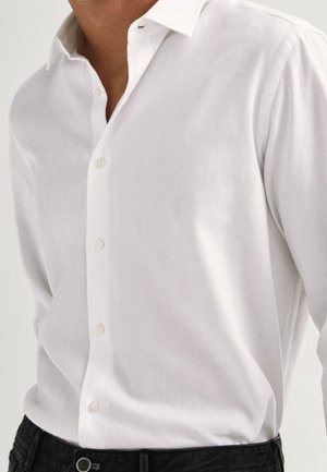 Homme portant une chemise blanche à manches longues boutonnée, rentrée dans un pantalon noir, montrant le col de la chemise et les boutons avant.