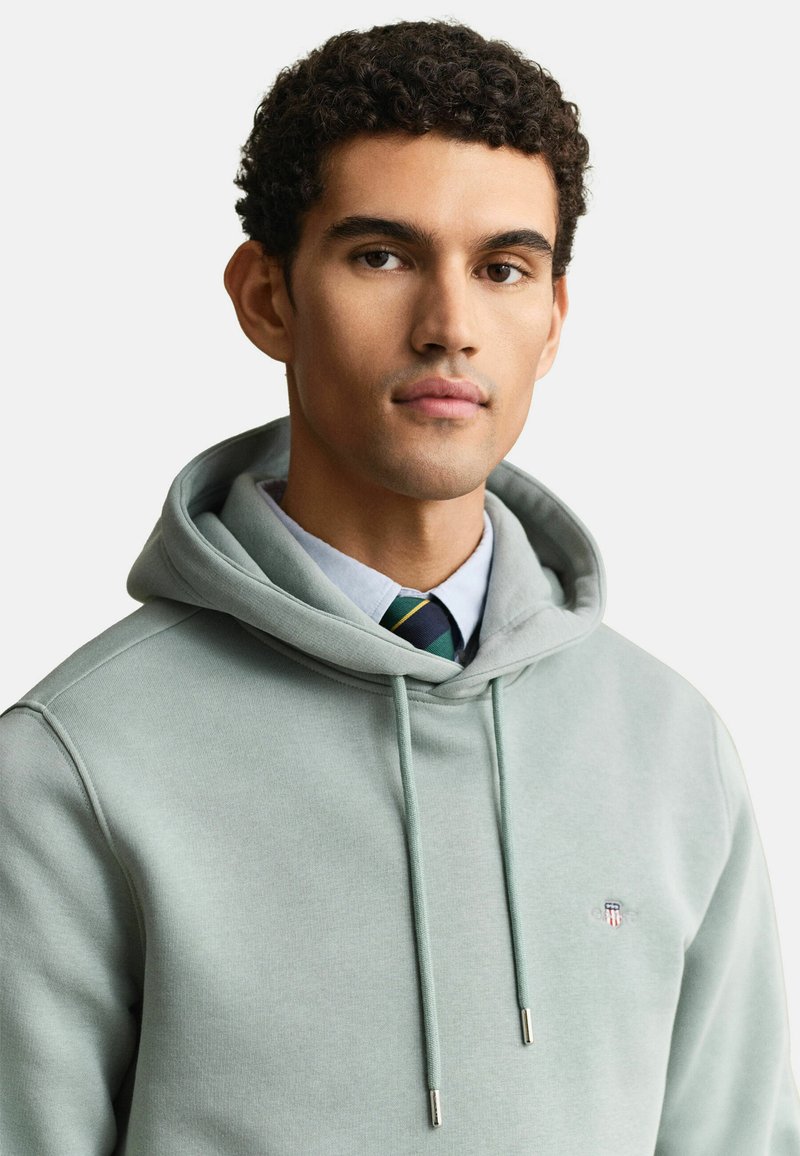 Giovane uomo con capelli ricci che indossa una felpa con cappuccio verde chiaro sopra una camicia con colletto e cravatta a righe, rivolto in avanti con un'espressione neutra.