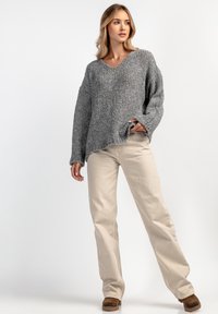 Szary dzianinowy sweter z dekoltem w serek, długimi rękawami i postrzępionym wykończeniem, w połączeniu z beżowymi spodniami typu wide-leg i brązowym obuwiem.