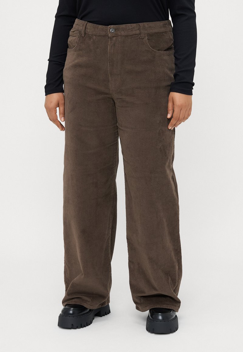 Corduroy brown pants with a wide-leg design, featuring front pockets, a button closure, and a soft texture. Worn with chunky black shoes.