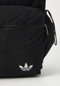 Rucsac negru texturat cu un buzunar cu fermoar, având un logo Adidas alb. Materialul are un model în formă de vafe și elemente de fixare rezistente.