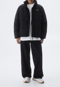 Doudoune noire à col haut avec fermeture éclair sur l'avant, présentant un petit logo et un design matelassé. Portée par-dessus une chemise blanche et un pantalon foncé.