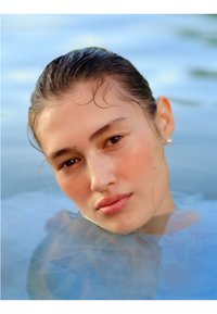 Gros plan d'une personne avec les cheveux mouillés et un teint lisse, partiellement immergée dans l'eau, avec un doux fond bleu et une boucle d'oreille en perle.