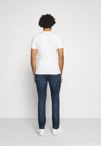 T-shirt bianca a maniche corte abbinata a jeans slim-fit blu scuro e sneakers bianche, mostrata da dietro su uno sfondo semplice.