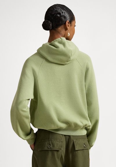 Camisola com capuz verde claro, de textura suave, com capuz e punhos canelados. Usada com calças verde-oliva que têm bolsos com botões.