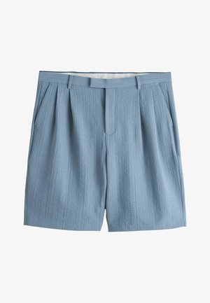 Nahaufnahme einer strukturierten blauen Hosentasche mit Knopf, Gürtelschlaufen und einem Teil eines weißen Hemdes und Arms sichtbar.