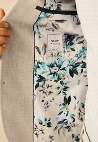Intérieur d'un blazer gris clair avec un motif floral en teal et crème. L'étiquette indique "NEXT TAILORING SLIM FIT". Texture satin lisse.