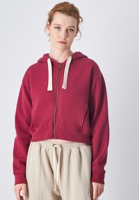 Sudadera corta burdeos con cremallera y capucha ajustable, cintura con cordón beige y puños acanalados. Tejido suave con costuras visibles.