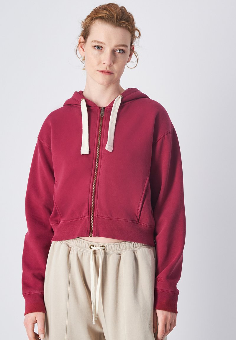 Sudadera corta burdeos con cremallera y capucha ajustable, cintura con cordón beige y puños acanalados. Tejido suave con costuras visibles.