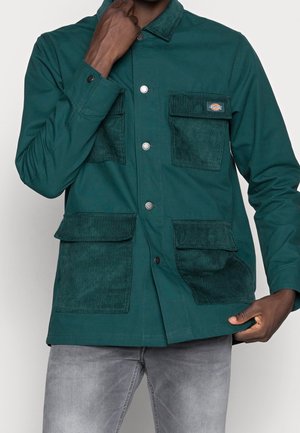 Homme portant une veste verte à fermeture boutonnière et quatre poches plaquées en velours côtelé, associée à un jean gris clair, main touchant le col.