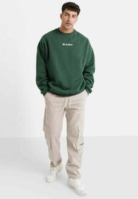 Oversized donkergroene sweatshirt met witte tekst, gecombineerd met beige cargobroek met zijzakken en drukknoopjes, gedragen met witte sneakers.