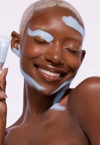 Crema facial en un tubo azul claro sostenido por una persona con cabello corto rubio. Acentos de crema azul sobre la piel, textura suave, tono de piel marrón cálido.