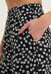 Svart bukser med et hvitt blomster mønster, elastisk midje, lett stoff, sidelomme uthevet av en hånd, og avslappet passform.