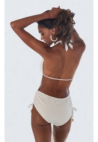 Femme à la peau foncée et aux cheveux bouclés portant un bikini blanc avec un bas taille haute texturé, tenant ses cheveux relevés sur un fond clair uni.