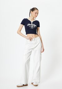 Camiseta corta azul marino con cuello blanco y gráfico de Brooklyn. Combinada con pantalones cargo blancos de pierna ancha que tienen bolsillos laterales. Zapatillas casuales.